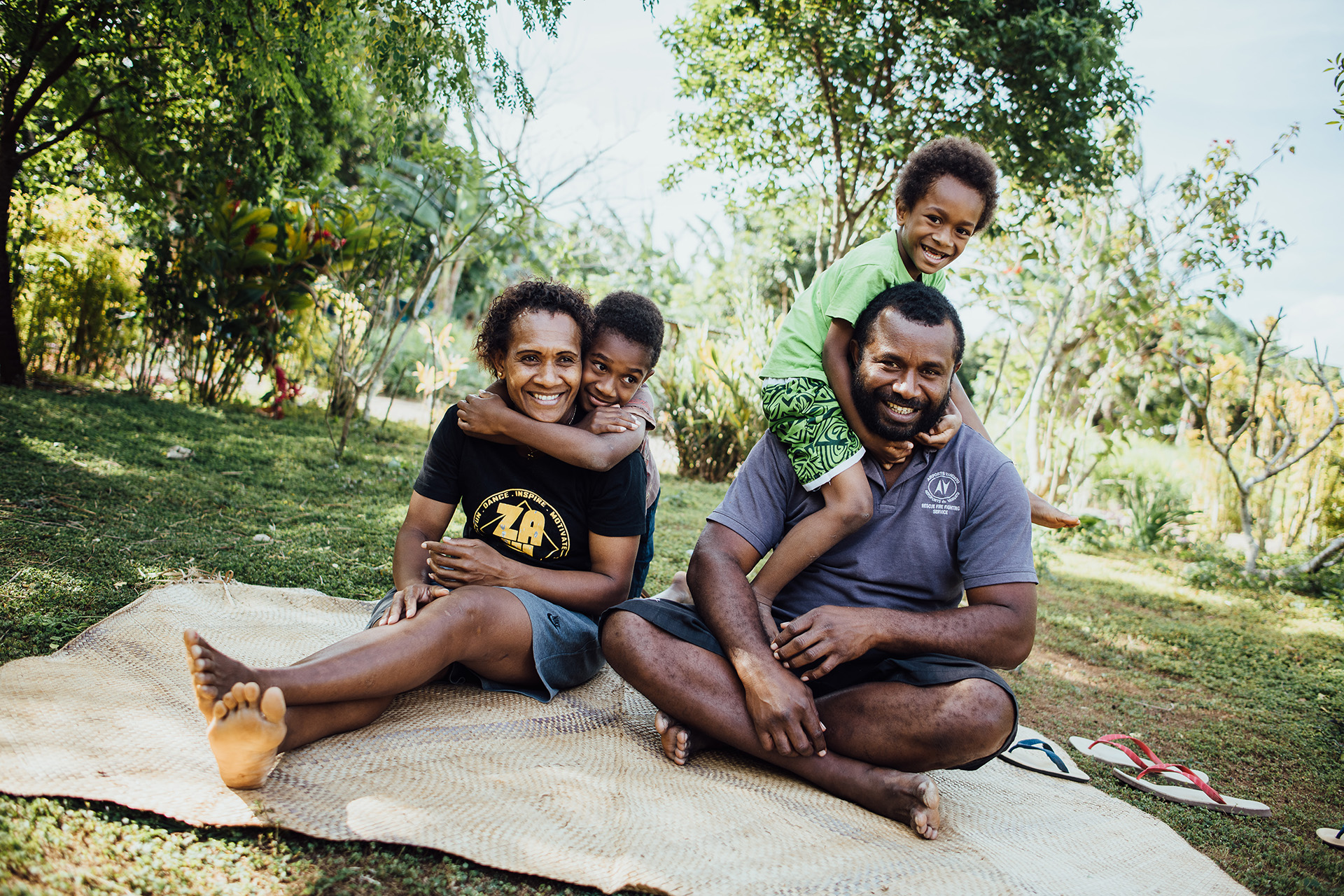 A family of four smiles while sitting on a mat in an outdoor grassy area. Anolyn Lulu is seated next to her partner, while two children hug them each from behind.